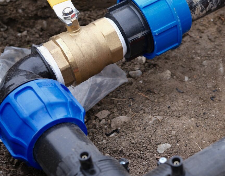 A brass ball valve with a yellow handle connects black underground pipes using blue compression fittings within a dirt trench - keatsagency.com
